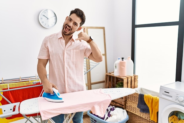 Jonge man met baard strijken kleren thuis glimlachend doend telefoongebaar met hand en vingers zoals praten aan de telefoon communicerende concepten