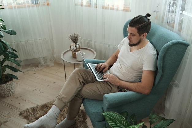 Jonge man met behulp van laptop typen en werken zittend op de fauteuil thuis