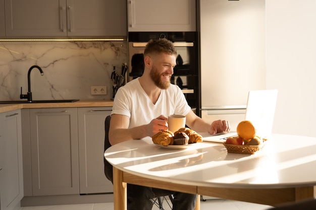 Jonge man met behulp van laptopcomputer en het drinken van thee of koffie. glimlachende europese bebaarde man zit aan tafel met heerlijk eten. interieur van keuken in modern appartement. zonnige ochtendtijd