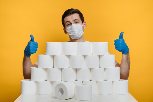 Jonge man met beschermend gezichtsmasker en medische handschoenen achter de stapel toiletpapier