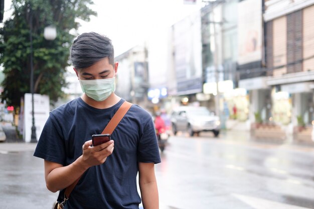 Jonge man met beschermend gezichtsmasker met behulp van mobiele telefoon in het centrum van de stad straat voorkomen dat virussen in de stad worden verspreid. Nieuw normaal concept.