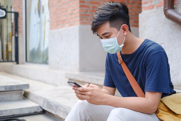 Jonge man met beschermend gezichtsmasker met behulp van mobiele telefoon in het centrum van de stad straat voorkomen dat virussen in de stad worden verspreid. Nieuw normaal concept.