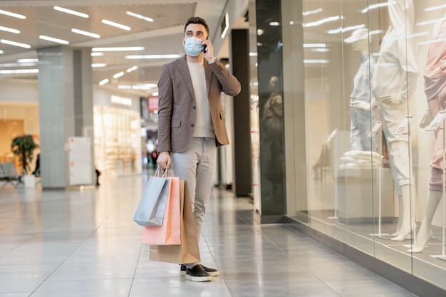 Jonge man met beschermend masker aan de telefoon tijdens het winkelen in het winkelcentrum tijdens pandemie