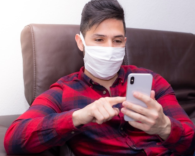 Jonge man met beschermend masker interactie met zijn smartphone binnenshuis voor quarantaine.