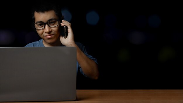 Jonge man met bril met behulp van een smartphone en computer laptop thuis, werk vanuit huis concept op zwarte achtergrond.