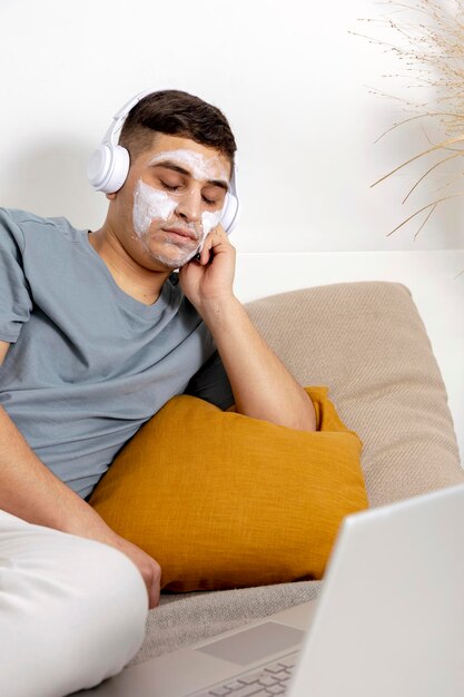 Jonge man met casual kleding en schoonheidsmasker op het gezicht liggend op bed met laptopcomputer en rusten Cosmetische voor mannen huidverzorging Man luisteren muziek of audioboek Ontspannen thuis