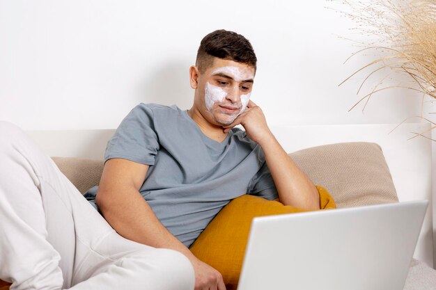 Jonge man met casual kleding en schoonheidsmasker op het gezicht liggend op bed met laptopcomputer en rusten Cosmetische voor mannen huidverzorging Man nieuws lezen op internet of film kijken Thuis ontspannen