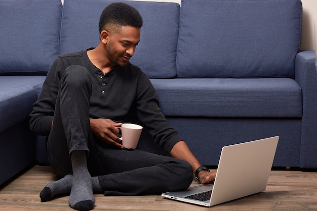 Jonge man met donkere huid die thuis met zijn laptop werkt, houdt kop met hete drank, zittend dichtbij bank op vloer