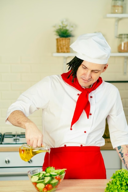 Jonge man met dreadlocks in uniform olijfolie gieten op groentesalade in de keuken Positieve chef-kok die salade voorbereidt en gezonde olijfolie uit de kan toevoegt