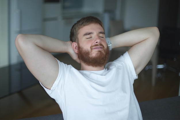 Jonge man met een baard alleen thuis ontspannen in de woonkamer op de bank zitten en mediteren in de