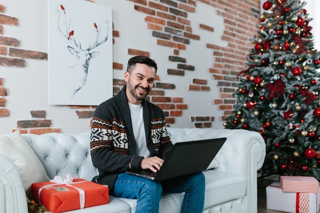 Jonge man met een baard die lacht en thuis op een laptop werkt, zittend op de bank tijdens de nieuwjaarsvakantie