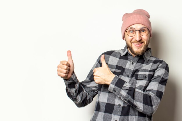 jonge man met een baard in een hoed, een geruit hemd maakt een gebaar duim omhoog op een afgelegen wit. Het gebaar is in orde, oké, geverifieerd