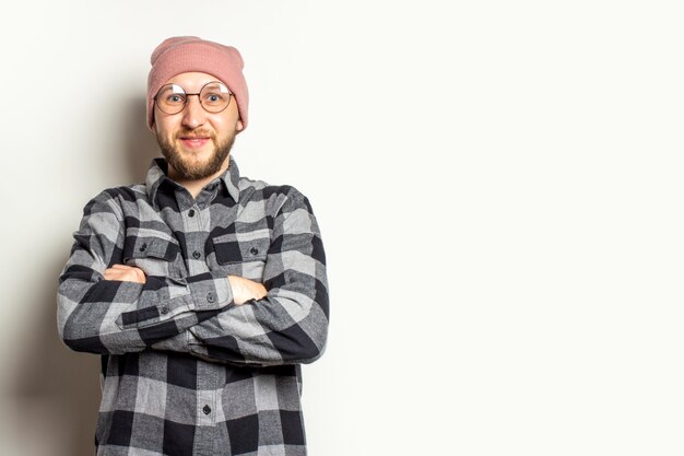 jonge man met een baard in een hoed, een geruite overhemd en een bril met armen gekruist op zijn borst op een afgelegen wit.