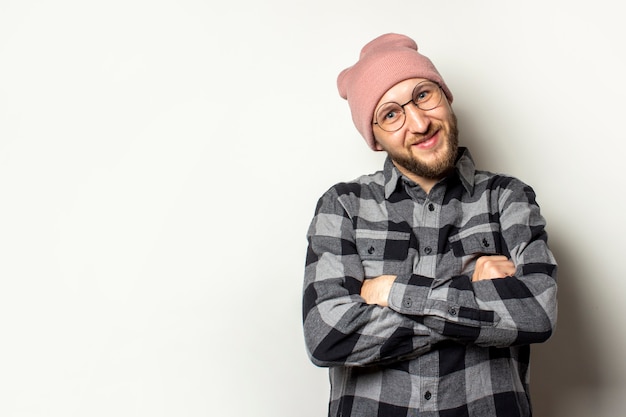 jonge man met een baard in een hoed, geruite overhemd en een bril met gekruiste armen op zijn borst lachend op een afgelegen wit.