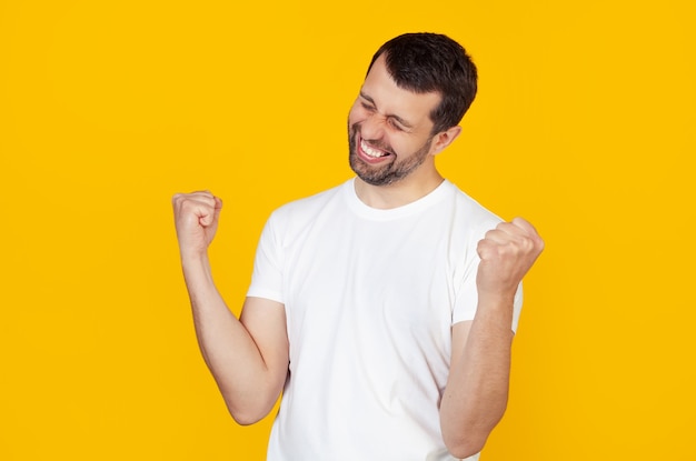 jonge man met een baard in een wit t-shirt is erg blij en opgewonden, een winnaar gebaar maken met opgeheven handen, glimlachen en schreeuwen succes Viering concept.