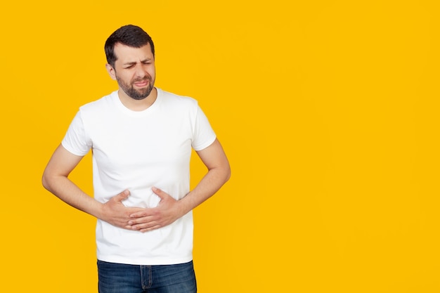 jonge man met een baard in een wit T-shirt met een hand op zijn buik vanwege indigestie, misselijkheid.