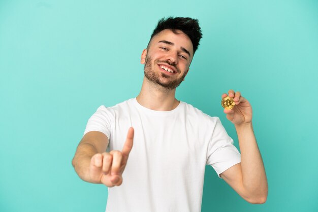 Jonge man met een Bitcoin geïsoleerd op een blauwe achtergrond die een vinger laat zien en optilt