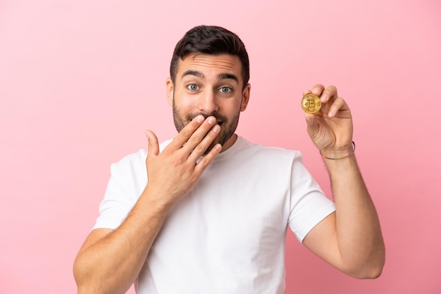 Jonge man met een Bitcoin geïsoleerd op een roze achtergrond, blij en glimlachend die de mond bedekt met de hand