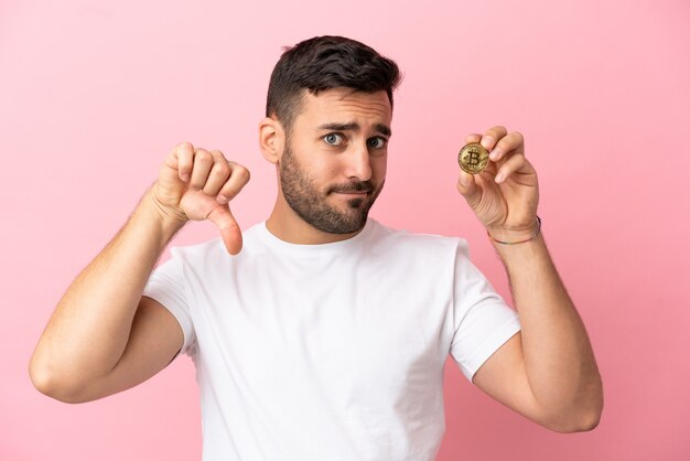 Jonge man met een Bitcoin geïsoleerd op een roze achtergrond met duim omlaag met negatieve uitdrukking