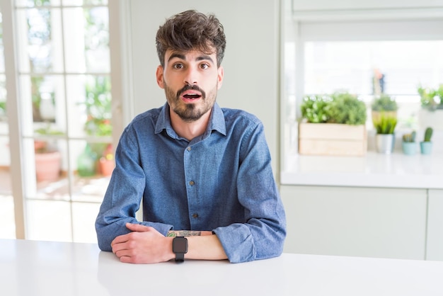 Jonge man met een casual shirt zittend op een witte tafel bang en geschokt van de angst van de verrassingsuitdrukking en het opgewonden gezicht