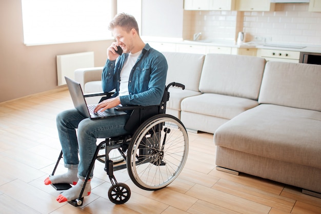 Jonge man met een handicap zittend op rolstoel. Bezig met laptop en praten over telefoon. Alleen in grote kamer met daglicht.