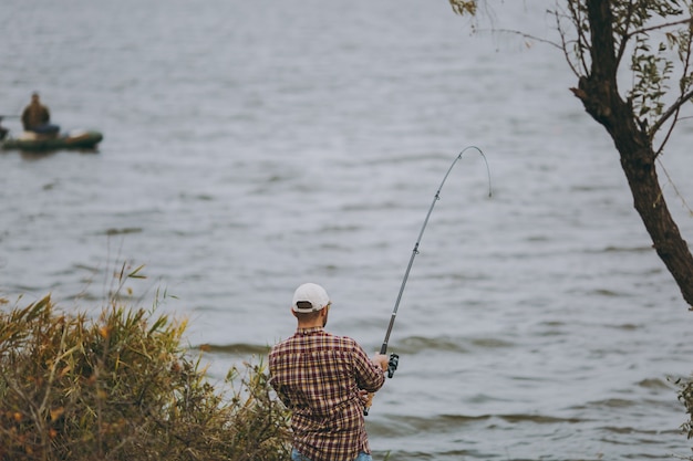 Jonge man met een hengel in geruit hemd en pet werpt een aas en vissen tegen de achtergrond van de boot op een meer vanaf de kust in de buurt van struiken en riet. Lifestyle, recreatie, vrije tijd concept.