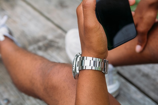 Jonge man met een horloge om zijn pols starend naar zijn telefoon terwijl hij in een park zit