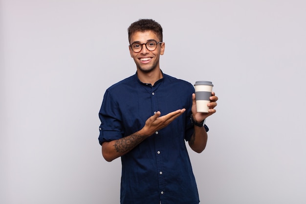 Jonge man met een koffie die vrolijk glimlacht, zich gelukkig voelt en een concept in exemplaarruimte met handpalm toont