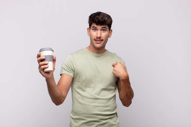 Jonge man met een kopje koffie, blij, verrast en trots, wijzend naar zichzelf met een opgewonden, verbaasde blik