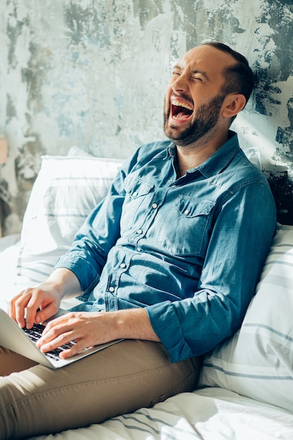 Jonge man met een laptop die op het bed zit en zijn ogen sluit terwijl hij lacht