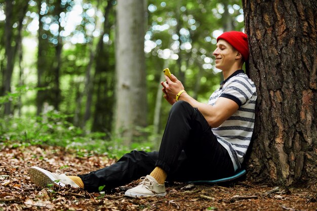 Jonge man met een rode hoed zittend aan de boom in het natuurbos tijdens trekkinghikingtraveling vrijheid lifestyle outdoor concept