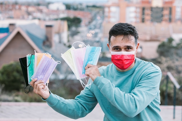 Jonge man met een rood chirurgisch masker met een handvol gekleurde chirurgische maskers met zijn hand. Covid-19 concept