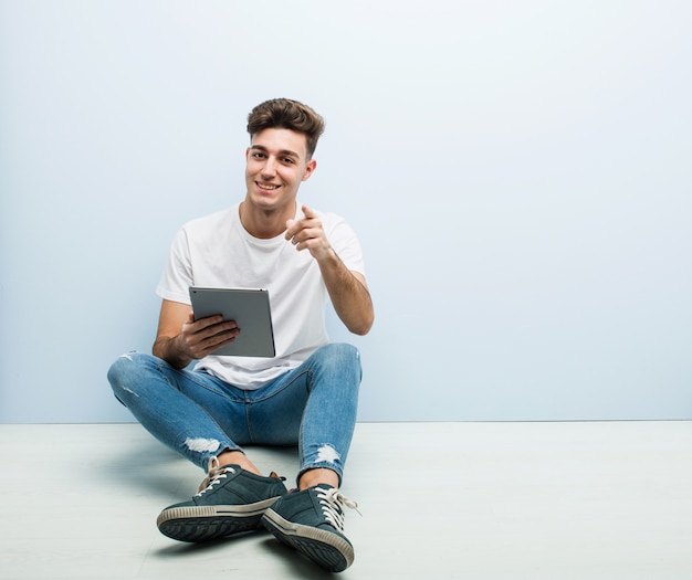 Jonge man met een tablet zit indoor vrolijke glimlach wijst naar voren.