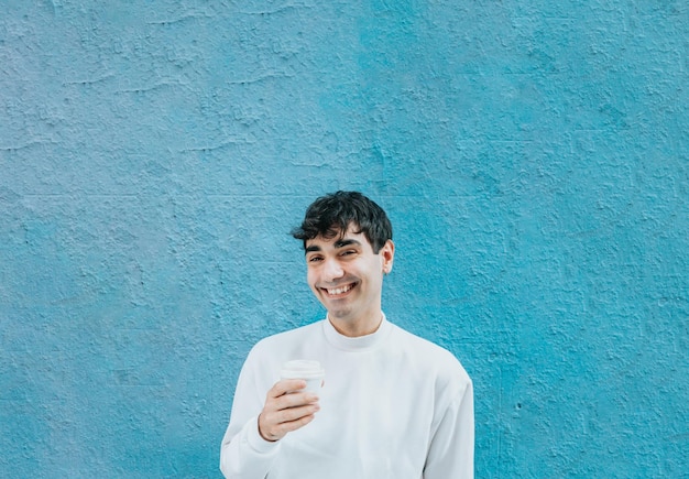 Jonge man met een take-away koffie over een grote blauwe muur terwijl hij lacht naar de camera. Modern levensstijlconcept. Witte, zoetere urban outfit. Madrid dagen.