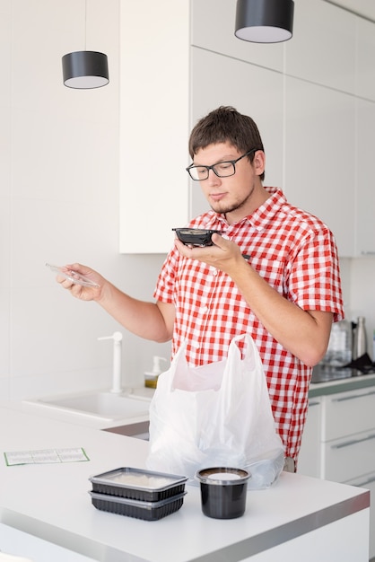 Jonge man met een wegwerp plastic zak met voedselbezorging in de moderne keuken, ruikend eten uit de doos