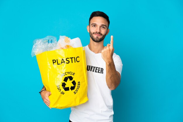 Jonge man met een zak vol plastic flessen om te recyclen geïsoleerd op blauwe muur doet komend gebaar