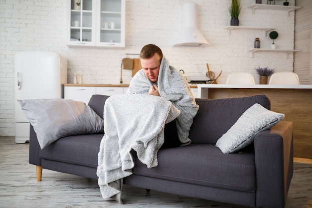 Jonge man met griep, zittend op de bank thuis, gewikkeld in een deken.