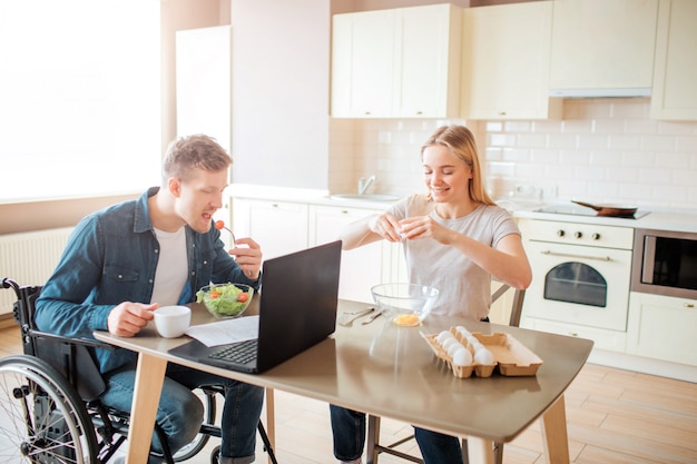 Jonge man met inclusiviteit en speciale behoeften salade eten in de keuken. Ga op een rolstoel zitten en studeer. De jonge vrouw zit bovendien en breekt eieren. Samenwerken.