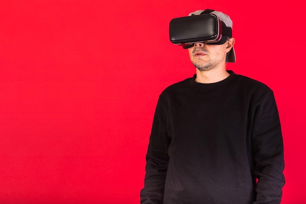 Jonge man met korte baard, gekleed in zwart sweatshirt, pet en virtual reality-bril verbaasd, op rode achtergrond. Technologie, VR, computing en hobby's concept.