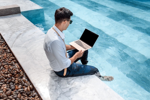 Jonge man met laptop zit bij het zwembad