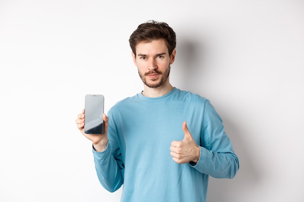 Jonge man met leeg smartphonescherm en duim omhoog, beveel mobiele applicatie of online winkelaanbieding aan, staande op een witte achtergrond