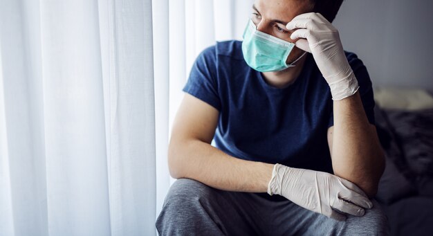 Jonge man met masker en handschoenen die door het raam kijken. blijf thuis, wees verantwoordelijk. alleen thuis.