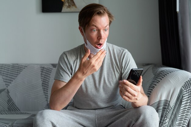 Jonge man met masker met behulp van telefoon en thuis in quarantaine geschokt kijken