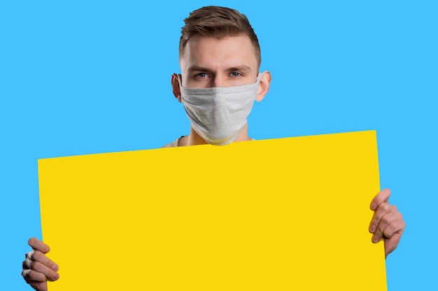 Foto jonge man met medisch masker geel papier poster tonen en camera kijken op blauwe achtergrond. reclame concept