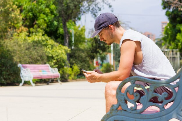 Jonge man met mobiele telefoon zittend op een bankje op een zonnige dag Jongen op mouwloos shirt en korte broek met behulp van slimme telefoon buitenshuis in de zomer
