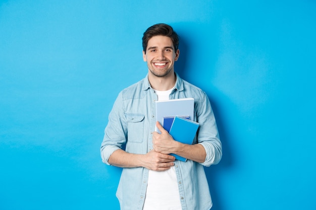 Jonge man met notitieboekjes en studiemateriaal, gelukkig glimlachend, staande over blauwe achtergrond