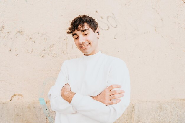 Jonge man met oortelefoons over een grote witte pastelmuur terwijl hij lacht naar de camera. modern levensstijlconcept. witte, zoetere urban outfit. madrid dagen. muziek leven