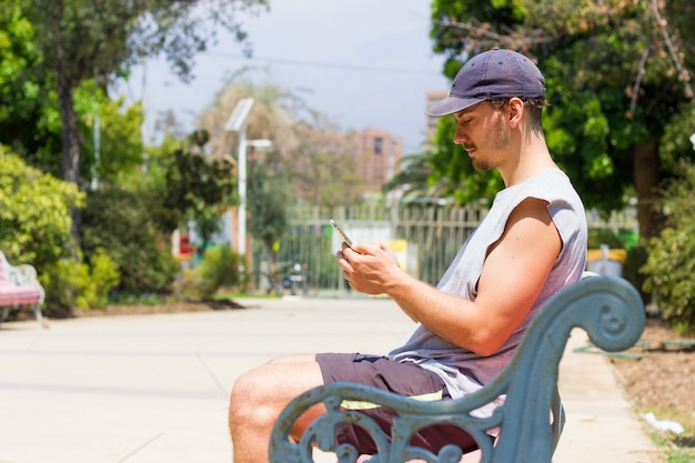 jonge man met pet met mobiele telefoon zittend op een bankje op een zonnige dag