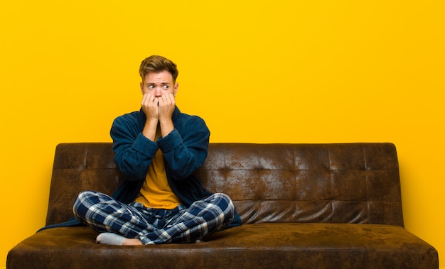 Jonge man met pyjama op zoek bezorgd, angstig, gestrest en bang, nagels bijten en op zoek naar laterale kopie ruimte. zittend op een bank