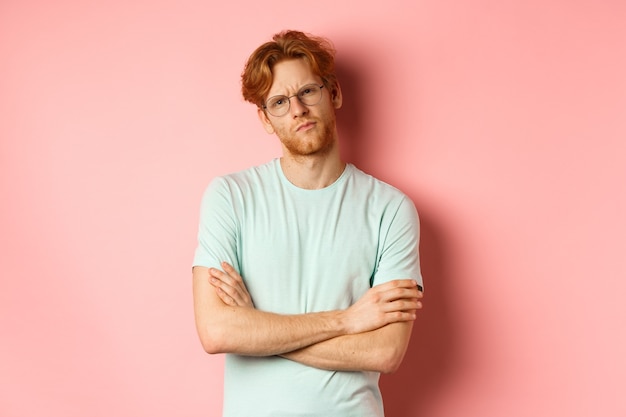 Jonge man met rood haar en baard, bril en t-shirt, armen gekruist op de borst, fronsend terwijl hij met een sceptische en twijfelachtige uitdrukking staarde, staande over roze achtergrond.